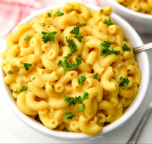 A close-up shot of a white bowl filled with creamy macaroni pasta, garnished with chopped green parsley, with a spoon resting in the bowl.